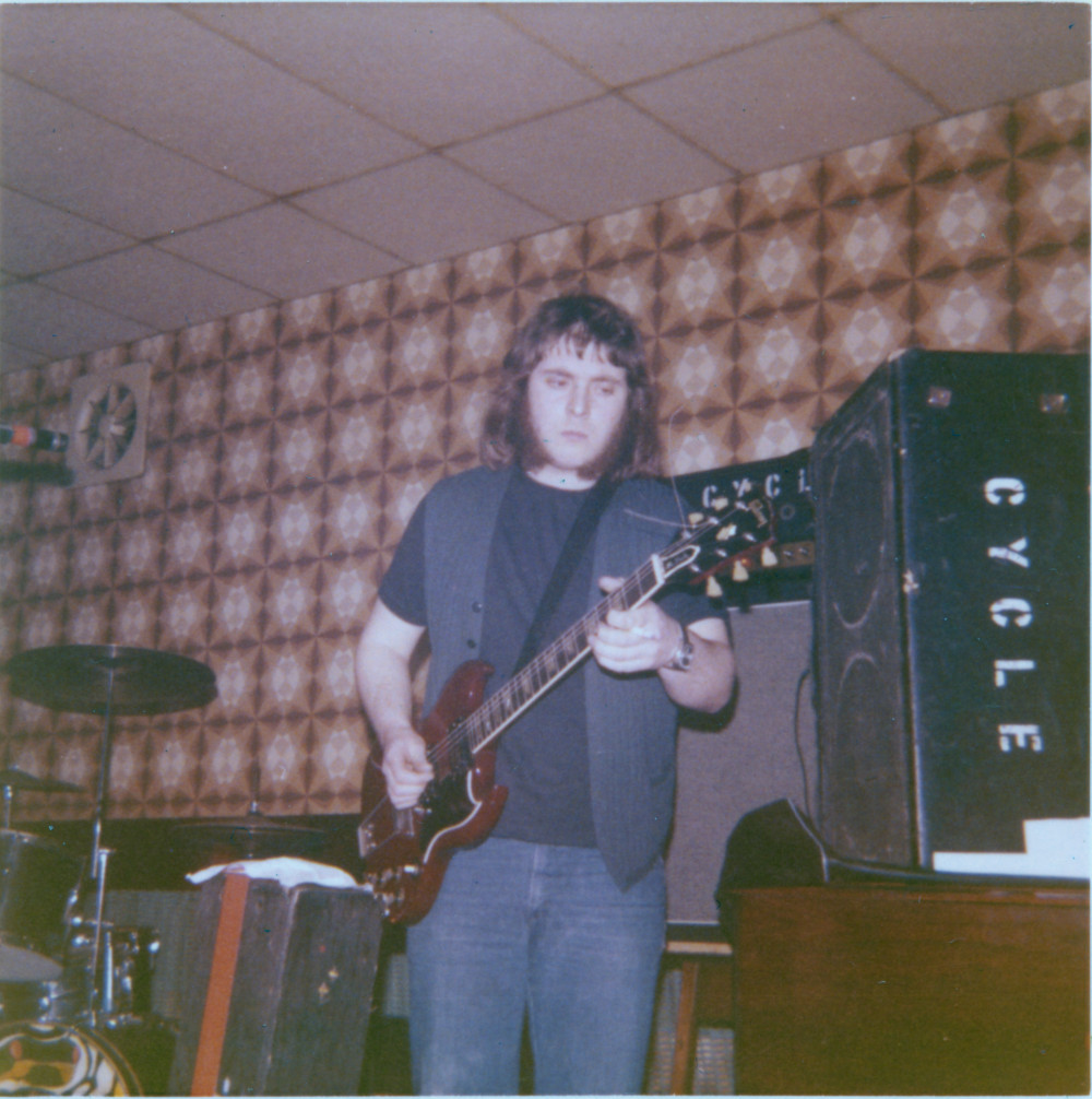John Whittingham playing guitar with a Marshall 100 Watt Super Bass amp behind him, and a PA monitor to his left