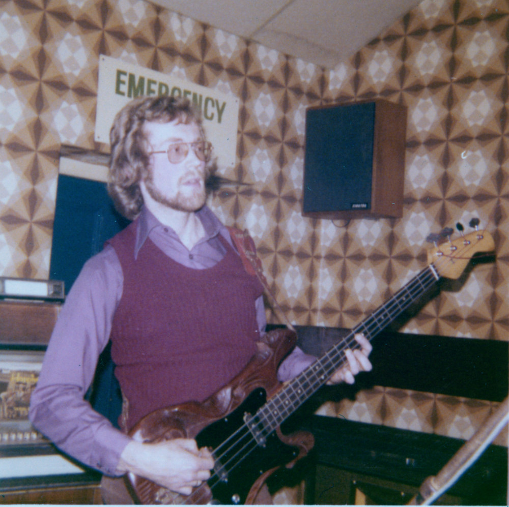 Cycle's bassist, Malcolm Harker, playing his custom carved bass guitar, an emergency exit sign is visible behind his head